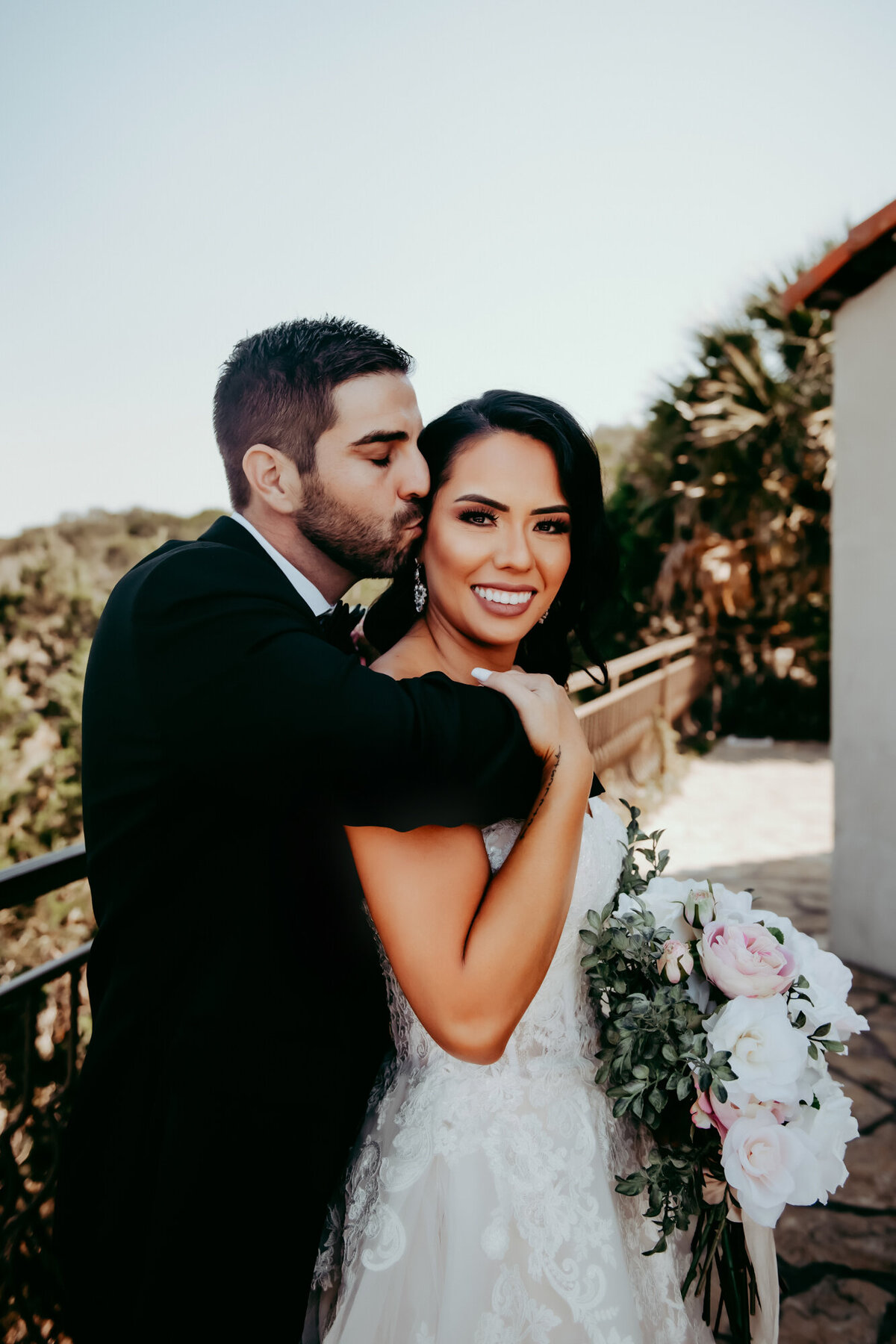 Couples Photographer,  groom embraces bride  and kisses her, she smiles