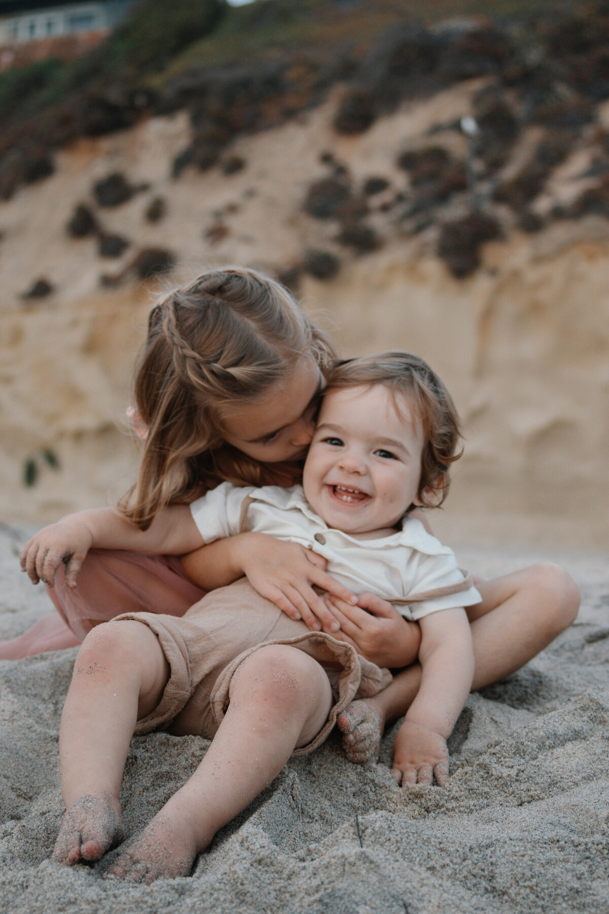 big sister hugs baby brother in the sand at the beach while baby brother laughs