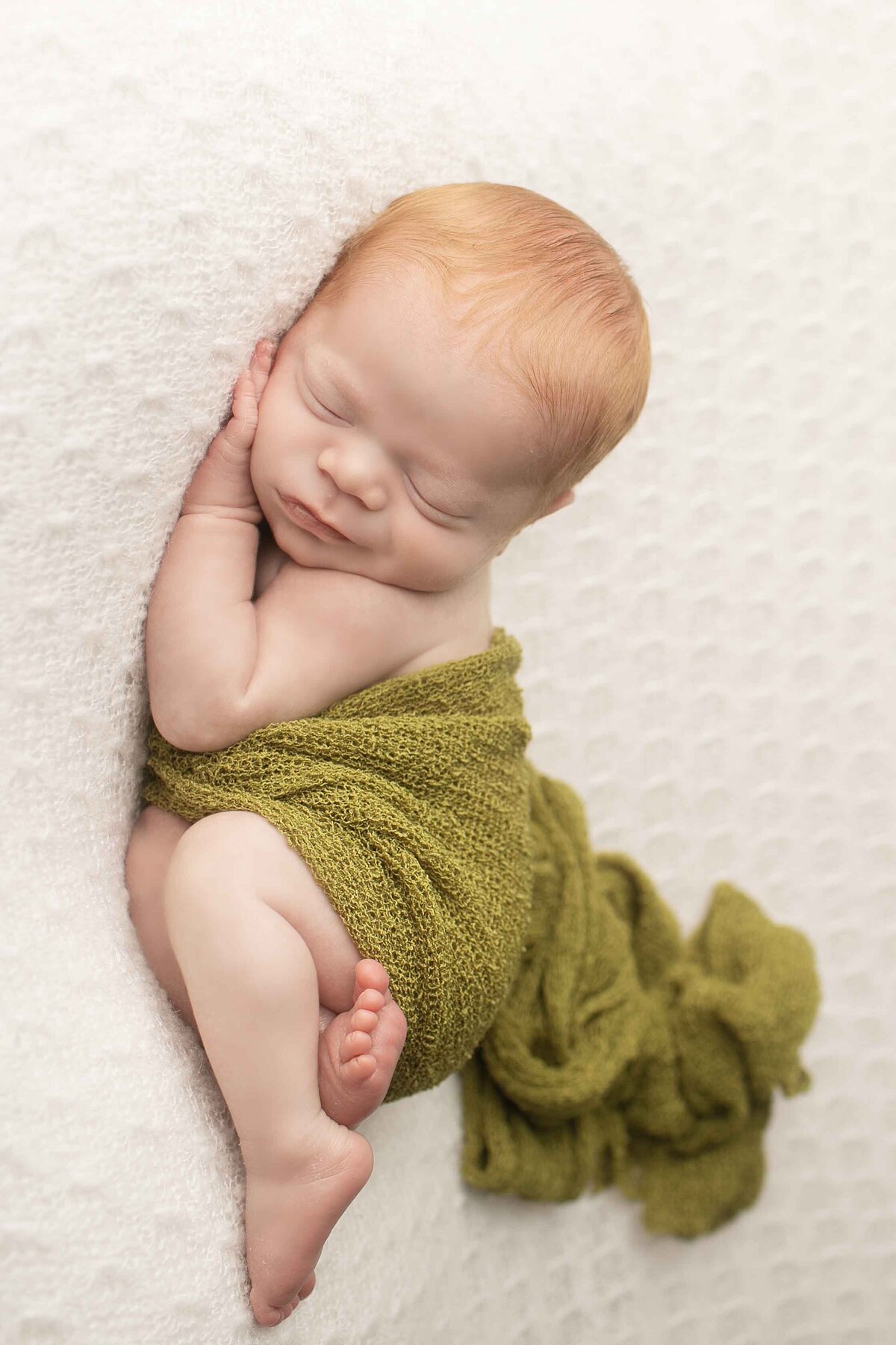 Newborn baby boy with striking red hair lying on his side on a textured backdrop, wrapped snugly for a cozy portrait.