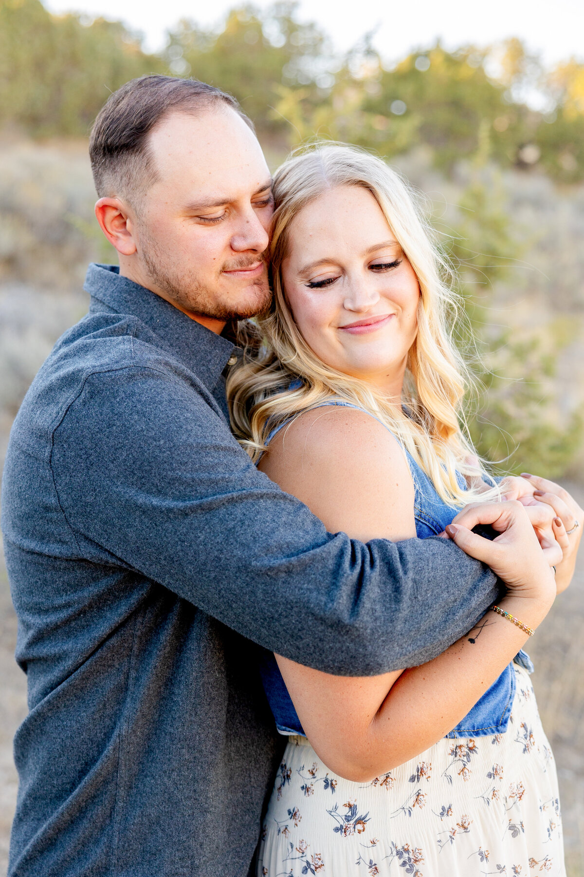 A couple stands close together, the man wrapping his arms around his wife as she leans into him with a soft smile, bathed in warm evening sunlight.
