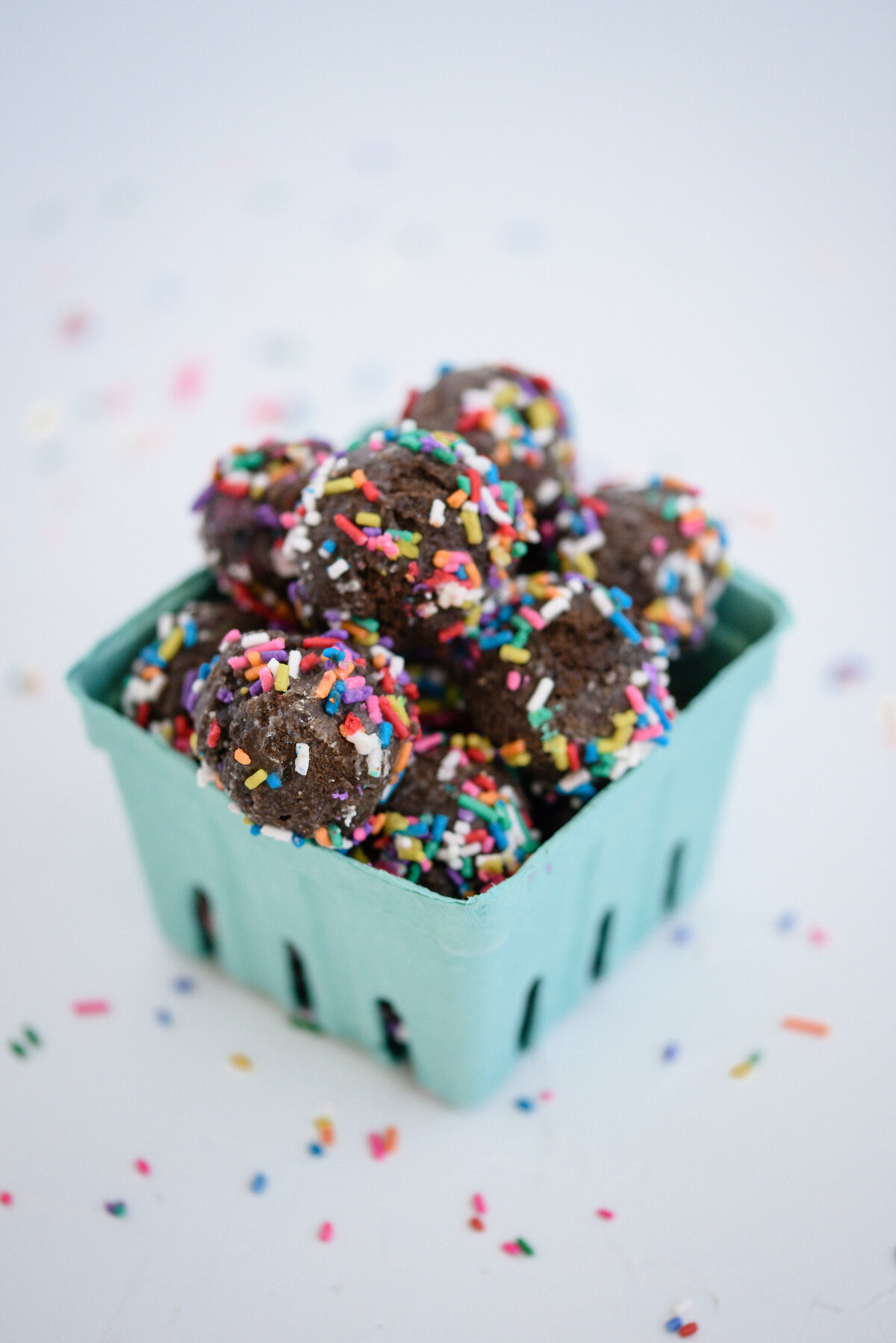 A product photographer near me takes a photo of a teal container holding chocolate donut holes with colored sprinkles all around