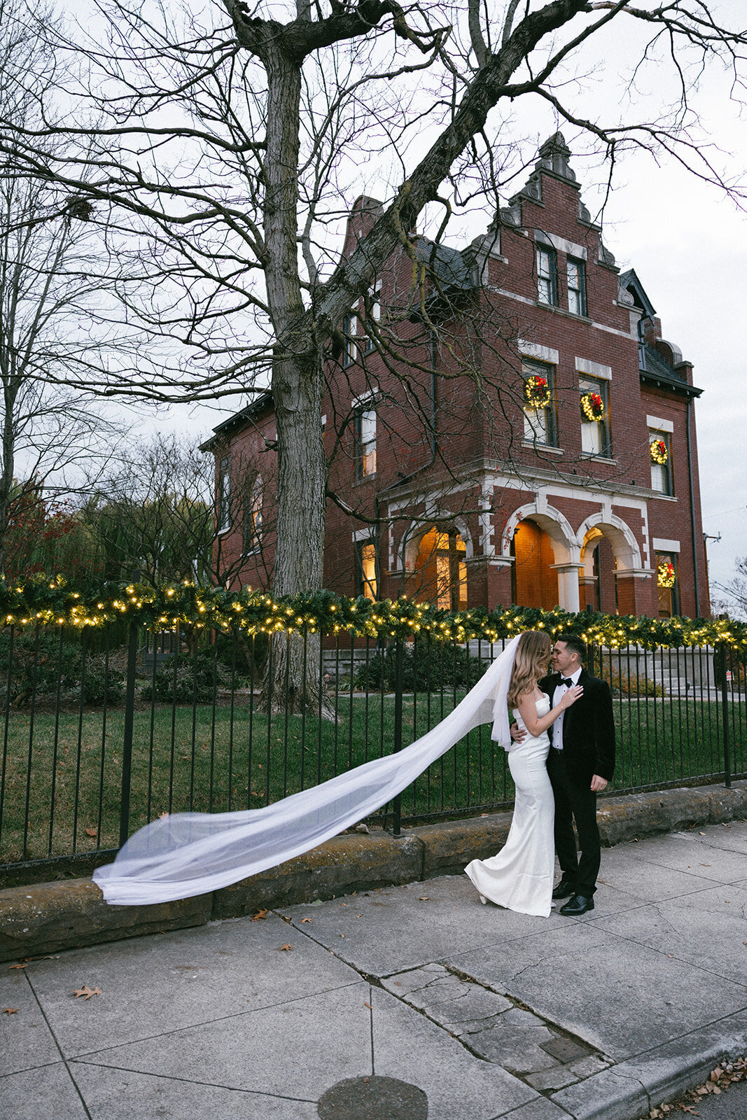 Brides veil dramatically flows in the wind as Christmas lights decorate the historic brick home behind them.
