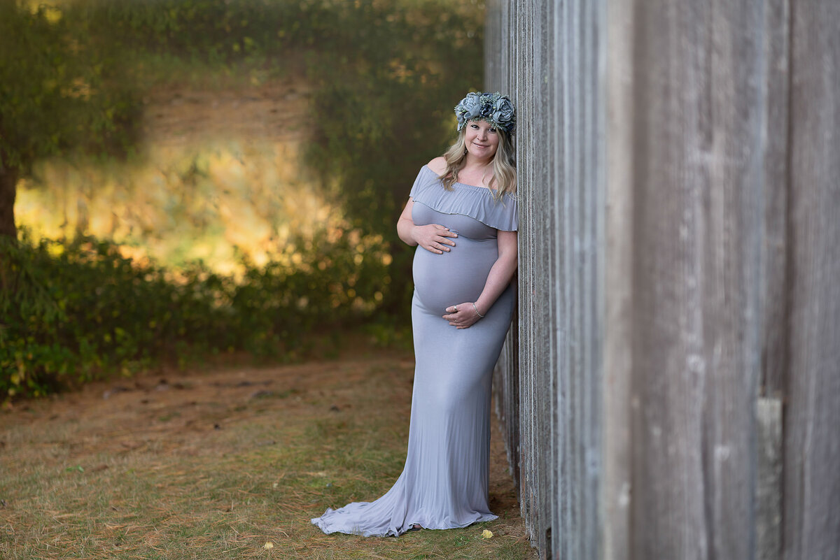 Expectant mom in lavender maternity gown is facing the camera and leaning against a rustic wood barn. She is wearing a flower crown. One hand us under her bump, the other atop of it. She is smiling at the camera with treed field behind her. Captured by Main Line photographer Liza Nicole.