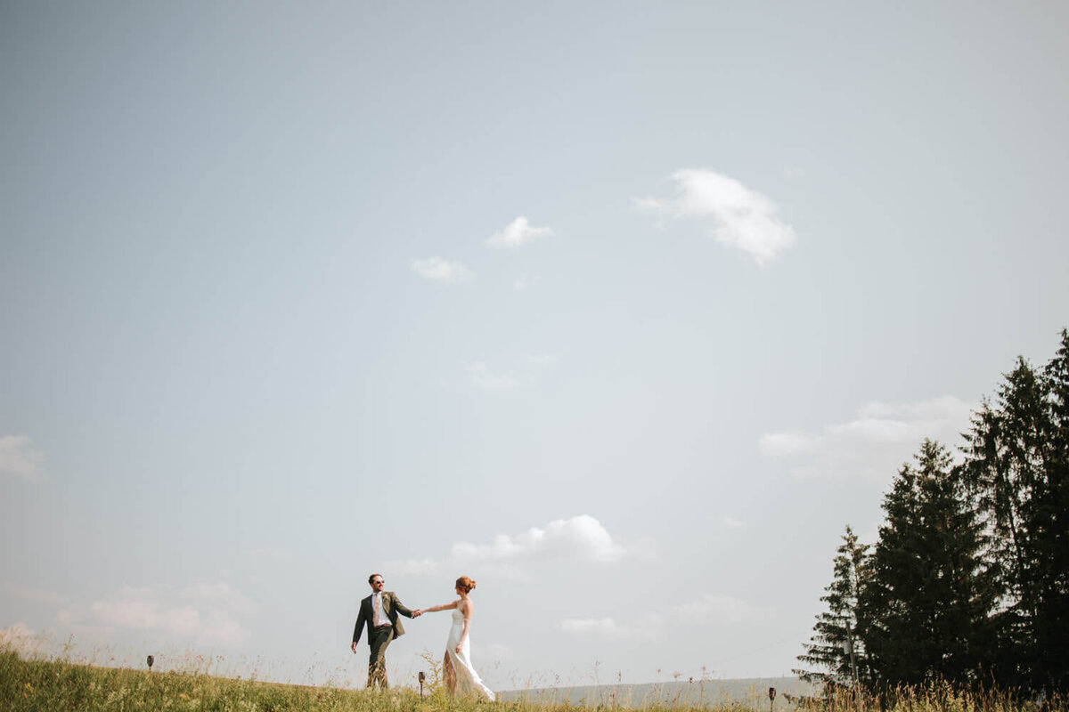 A bride and groom hold hands while walking on a hilltop under a clear sky.