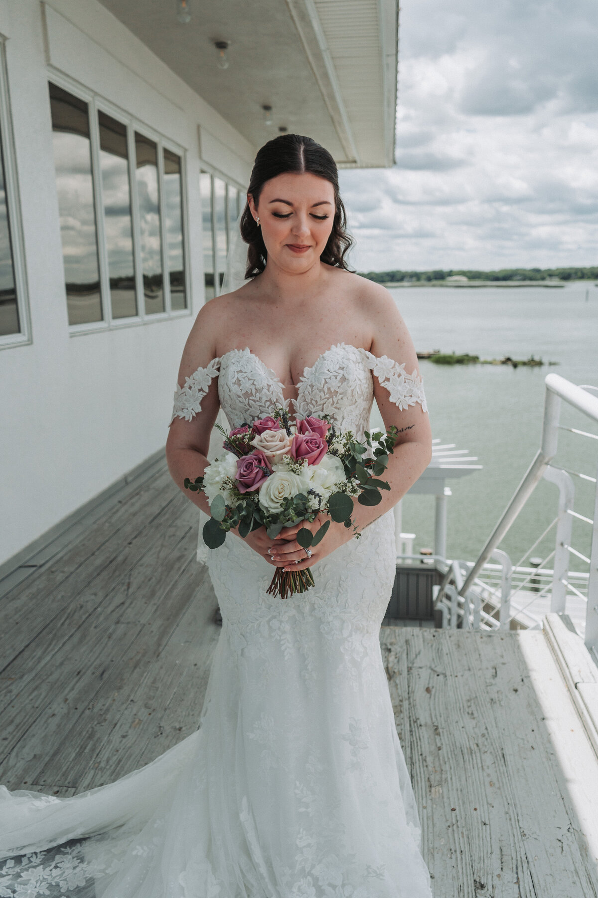 A stunning bridal portrait of a bride in her lace wedding gown, holding a pink and white floral bouquet, gazing down contentedly on the waterfront balcony of the Lesner Inn in Virginia Beach, captured by Quinn Photography LLC.