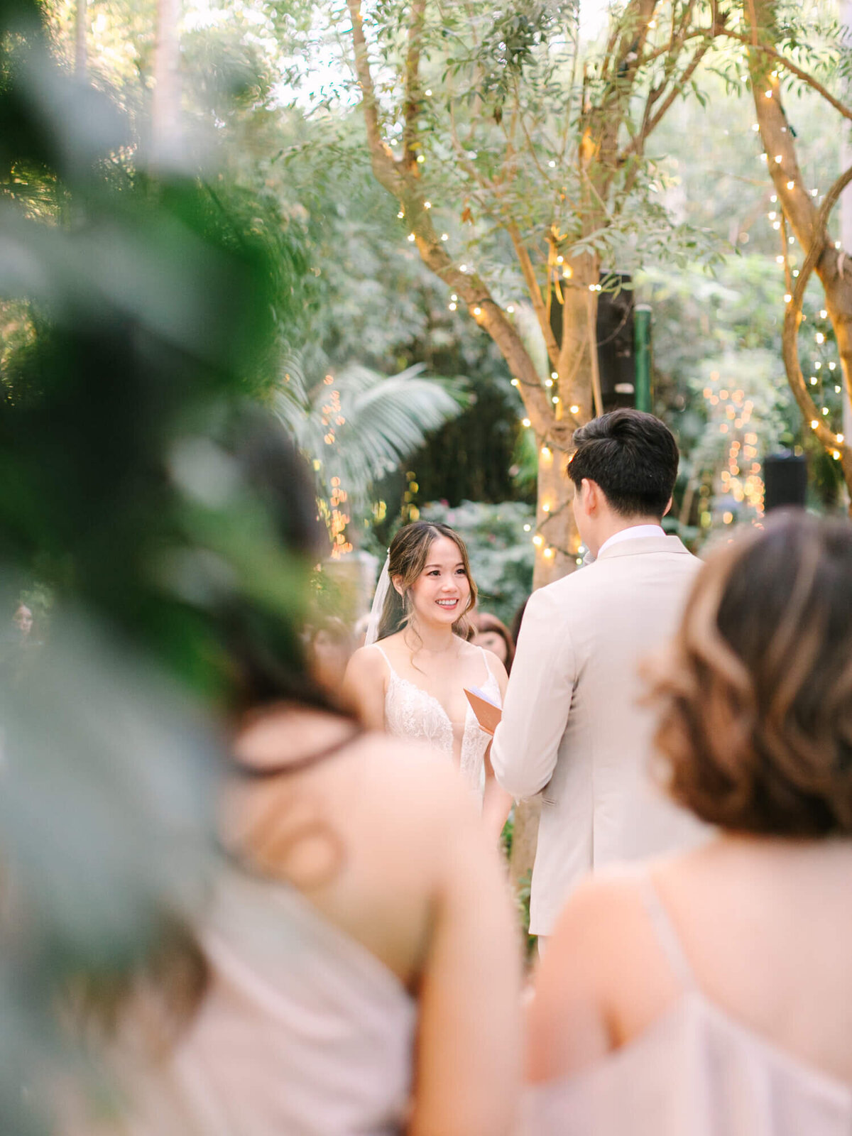 A bride and groom stand facing each other in a lush garden adorned with string lights.