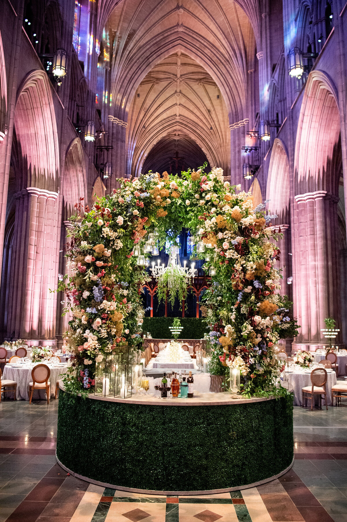 This photo was taken at Washington National Cathedral in Washington D.C. by photographer Twah Dougherty. The venue features towering arches and an elaborate floral arch centerpiece with vibrant flowers and greenery. Elegant tables with white linens complete the luxurious setting. Additionally, Current Films are the videographers for this event capturing all the beautiful moments on film.