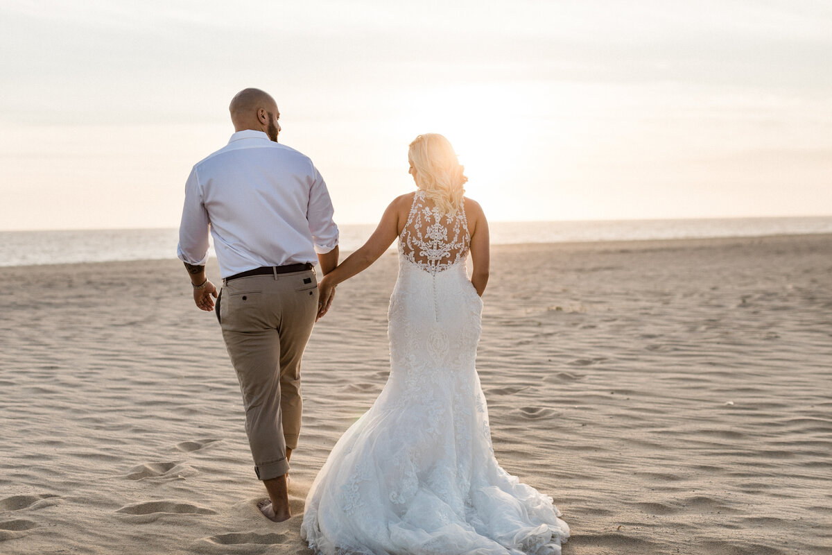 A bride and groom walk hand in hand along the beach at sunset. The bride is wearing a lace-back wedding gown, and the groom is in a white shirt and light-colored pants. Keywords: luxury destination weddings, beach wedding, destination wedding planning.