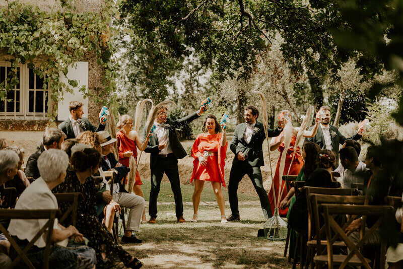 Amis des mariés faisant une danse face aux invités assis sur des chaises en bois dans un décor verdoyant. Séance photo mariage en vendée.