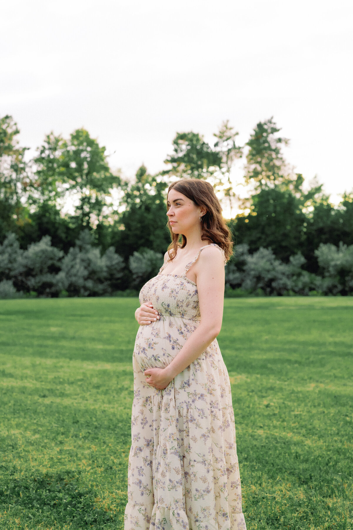 Mother standing in field cradling baby bump in floral dress