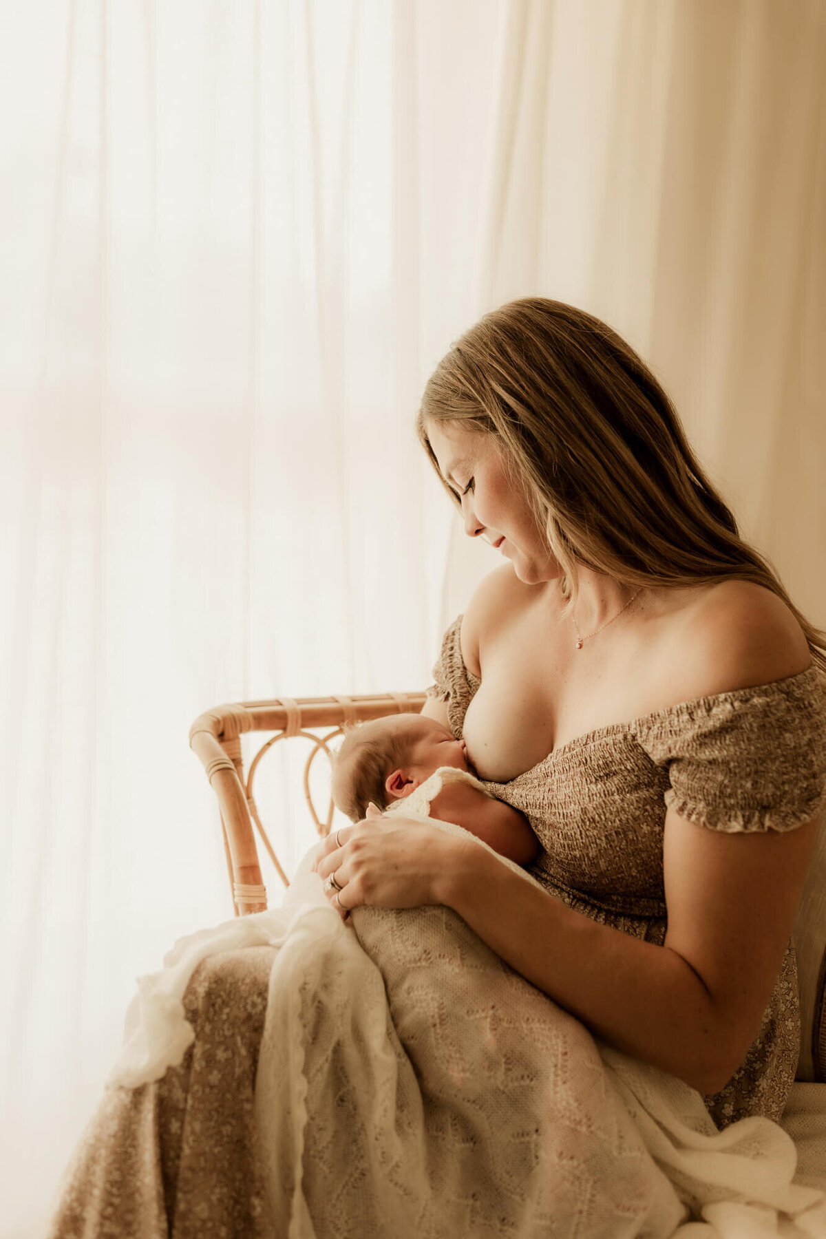 Mother breastfeeds her baby girl while sitting down on a rattan daybed near a window in OKC.