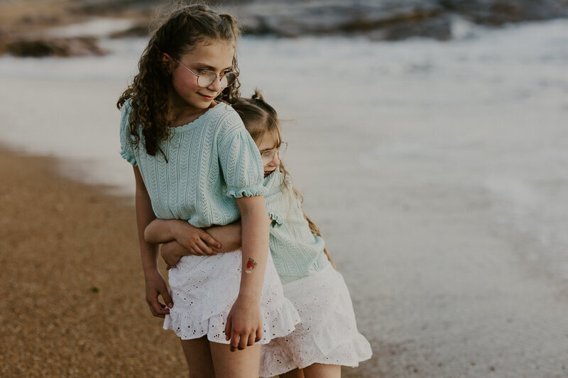 Petite fille enlaçant sa grande soeur de dos, toutes deux habillées en vert et blanc lors d'un shooting photo à la plage en Vendée.