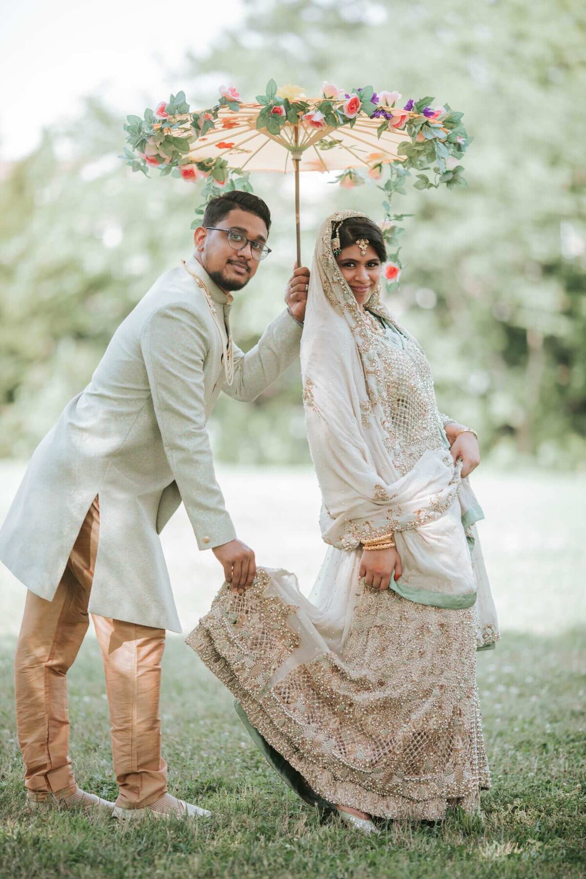 South Asian Groom holds an umbrella and the bride's dress. They are both surrounded by immense greenery.