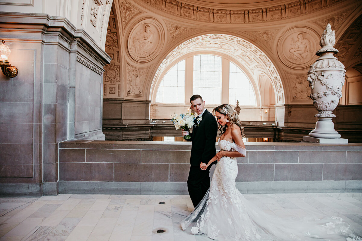 Bride and groom walking hand-in-hand on the 4th floor of San Francisco City Hall beneath the arched windows, captured by San Francisco elopement photographer Kristin Smith.