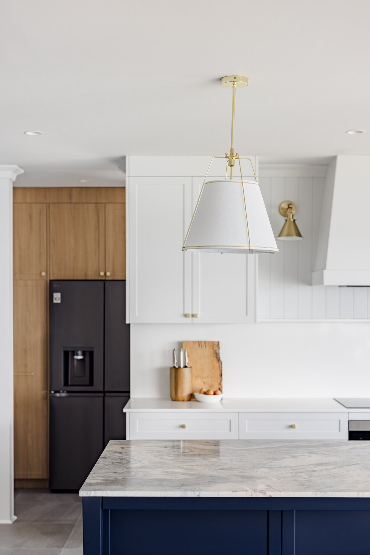 Modern kitchen featuring a marble island and navy base, complemented by a black fridge and white cabinets under a stylish pendant lamp. Crafted with bespoke interior design in mind, this Newcastle creation showcases the expertise of a skilled interior designer.