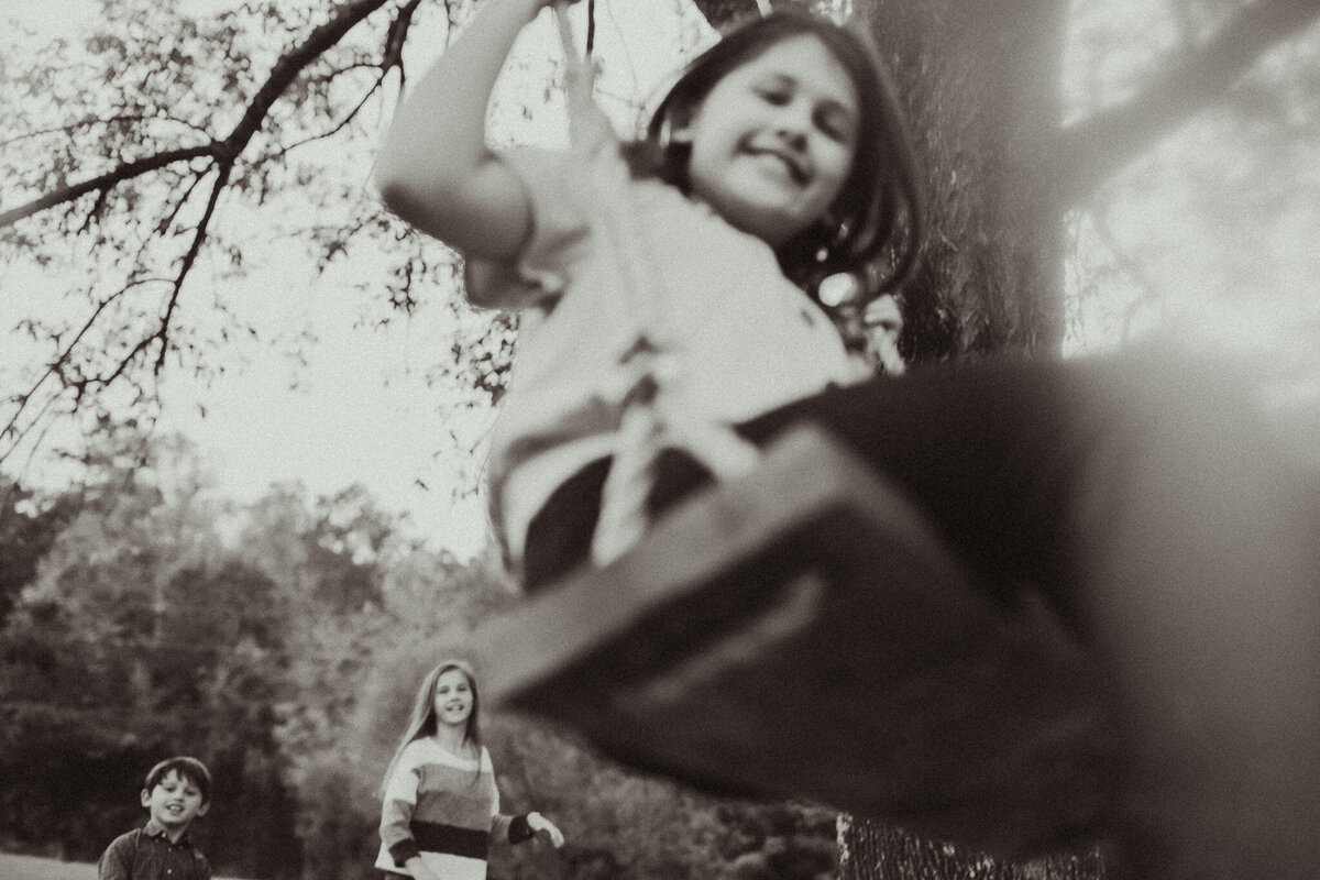 A young girl swinging while her mother and brother watch from a distance. 
