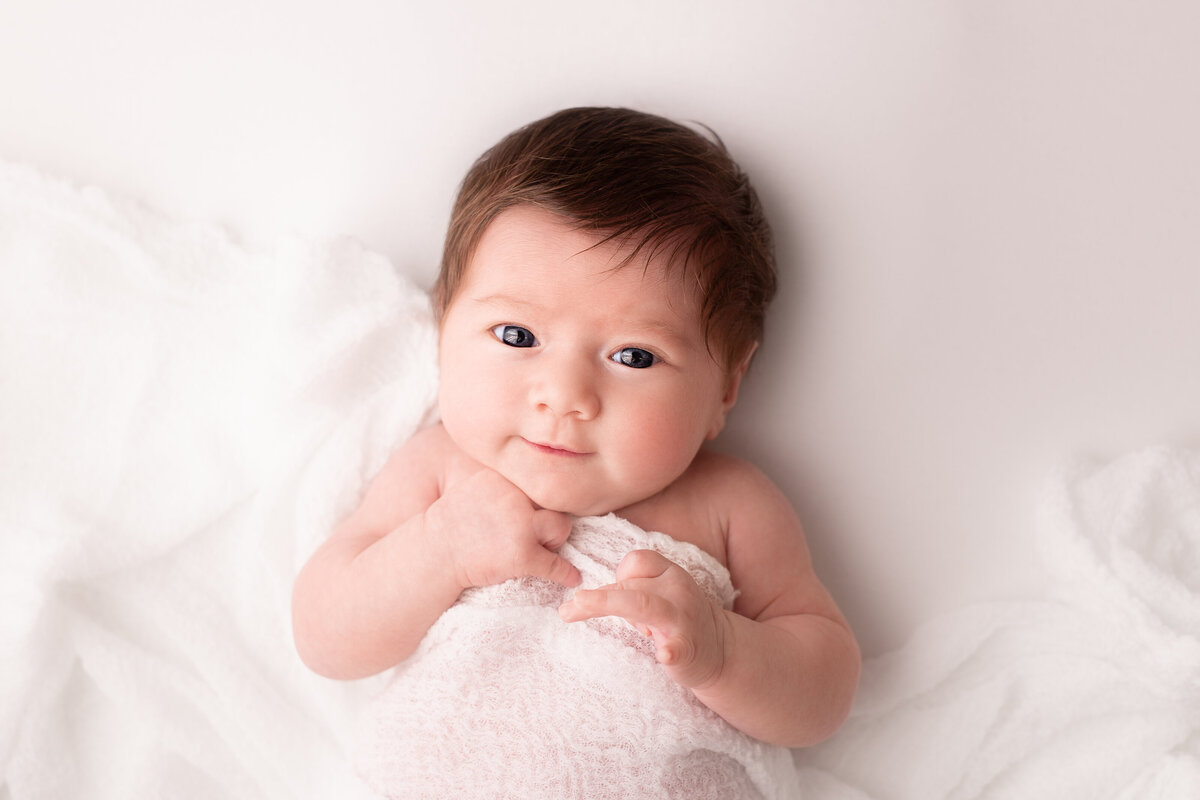 Baby lying on her back looking at the camera on a white background with white fabric over her.
