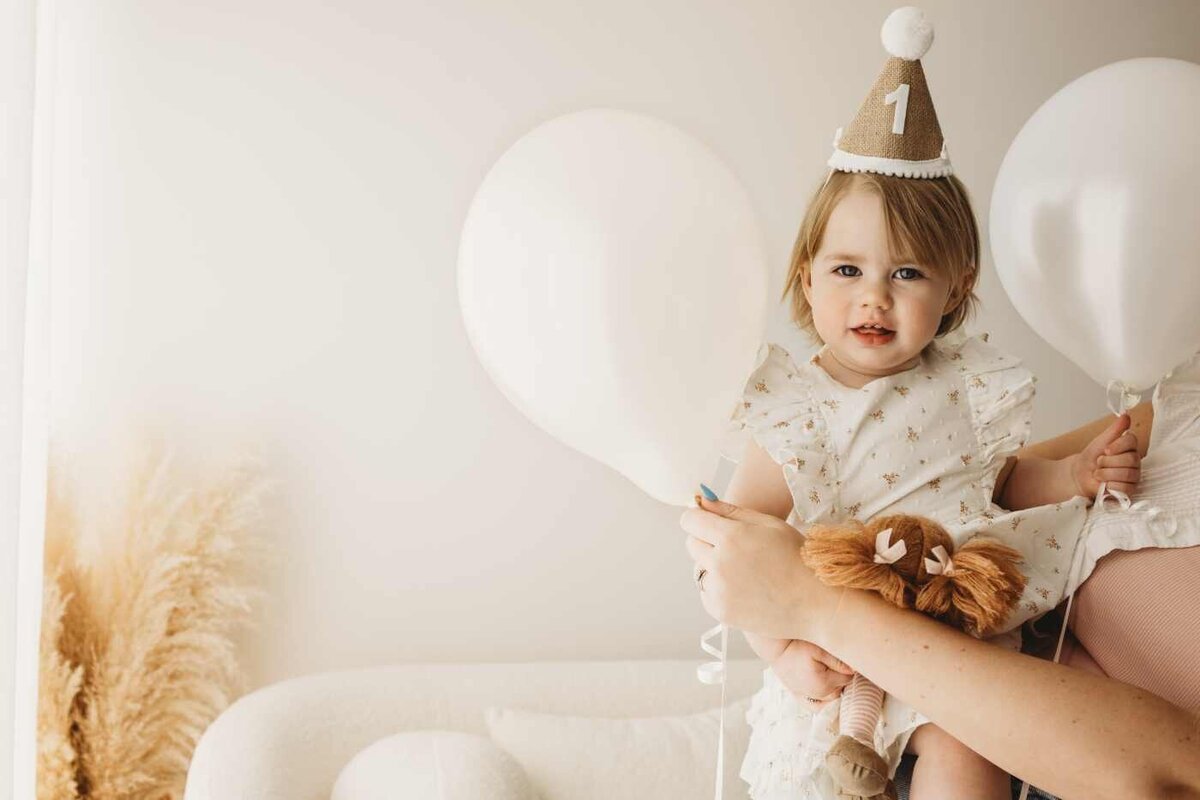 Ruua Hamdan Photography (UK): A baby in a floral dress is being held and is next to two big white balloons. The baby is wearing a party hat with the number 1 on it.