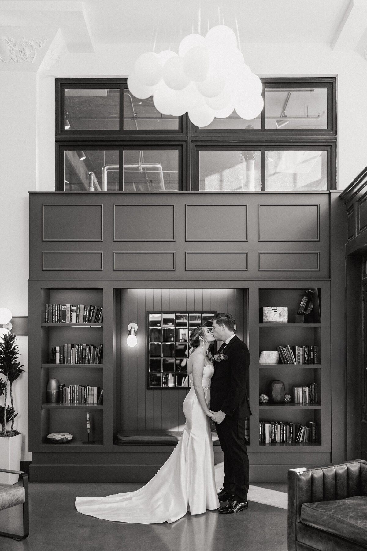 A black and white photograph of Cheyenne and Adam on their wedding day on New Year’s Eve in San Antonio, Texas. The bride and groom have just seen each other for the first time on their wedding day and they are facing each other, holding each other's hands at their sides and kissing. They are inside a building with a dark bookcase behind them and a chandelier of softly lit bulbs overhead. Wedding photography by Stacie McChesney/Vitae Weddings.