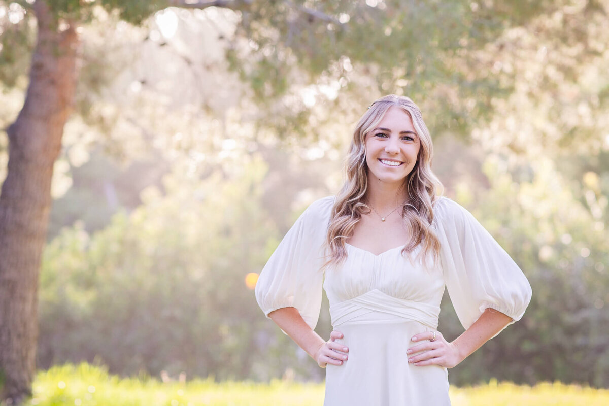 A woman in a white dress stands outdoors with hands on hips, smiling. The background is sunlit with greenery and trees, creating a soft, natural setting.