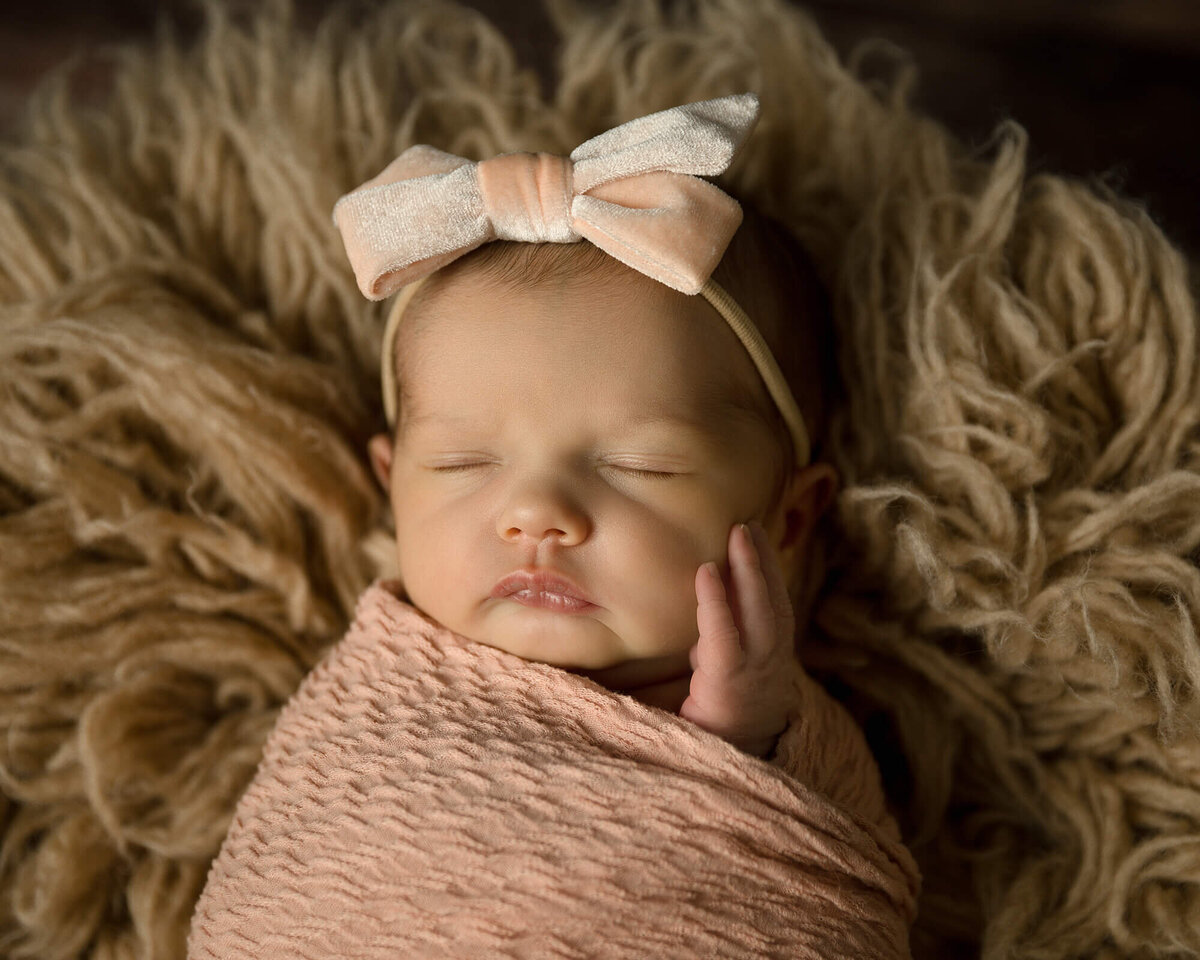 Newborn girl in a peach wrap and large bow headband.  She has her hand touching her cheek as she sleeps on a tan colored rug.