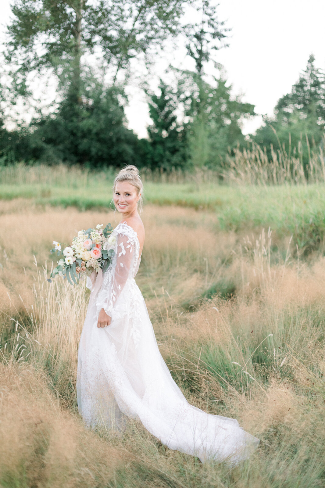 Stunning bridal inspiration; bride in wedding gown with long embroidered lace sleeves, standing in a field holding bouquet, captured by Ana Douglas Photography, timeless and authentic wedding photographer in Vancouver, BC. Featured on the Bronte Bride Vendor Guide.