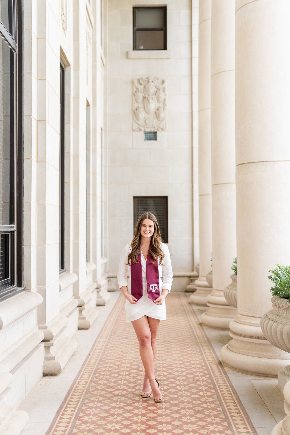 Texas A&M senior girl standing and holding stole and smiling with wearing a white dress in the columns of the Administration Building