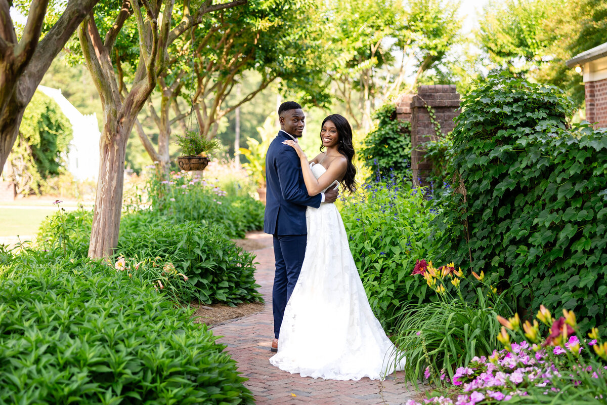 Immerse yourself in the beauty of love with this enchanting photo of a bride and groom in a lush garden, surrounded by trees, ivy, and vibrant flowers, capturing a moment of pure romance.
