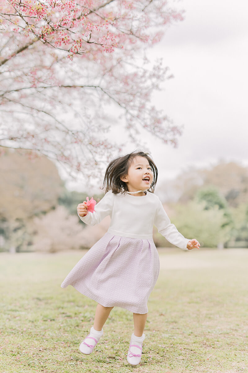 桜をバックに、3歳の女の子がお花を手に持ってジャンプしている躍動感のある写真