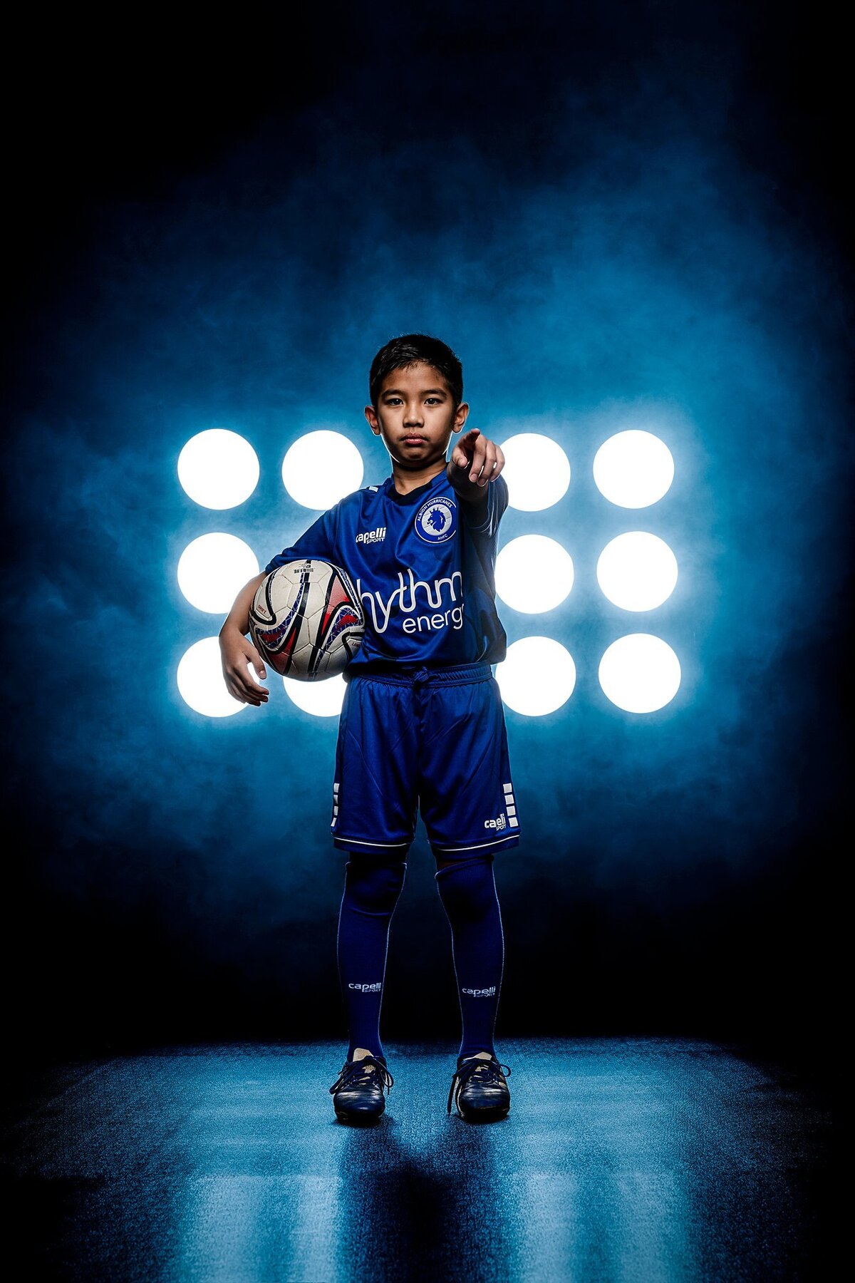 In the picture, a young man stands confidently in a blue soccer uniform, embodying the spirit of determination and athleticism. He holds a soccer ball securely tucked under his right arm, showcasing his passion for the sport. The backdrop features a striking blue smoky haze that adds a dramatic flair, enhancing the overall atmosphere of the image. His left arm is extended towards the camera, as if inviting viewers to join him on the field or to be part of the action. The two Pro Mod stadium lights positioned in front of him illuminate his focused expression, highlighting his game face—a look that reflects intensity and readiness. This carefully curated studio setting combines elements of sports photography and artistic flair, creating a compelling visual story of ambition and dedication. The blend of colors and lighting draws attention to the young athlete, making it clear that he is prepared to take on any challenge that comes his way on the soccer field.