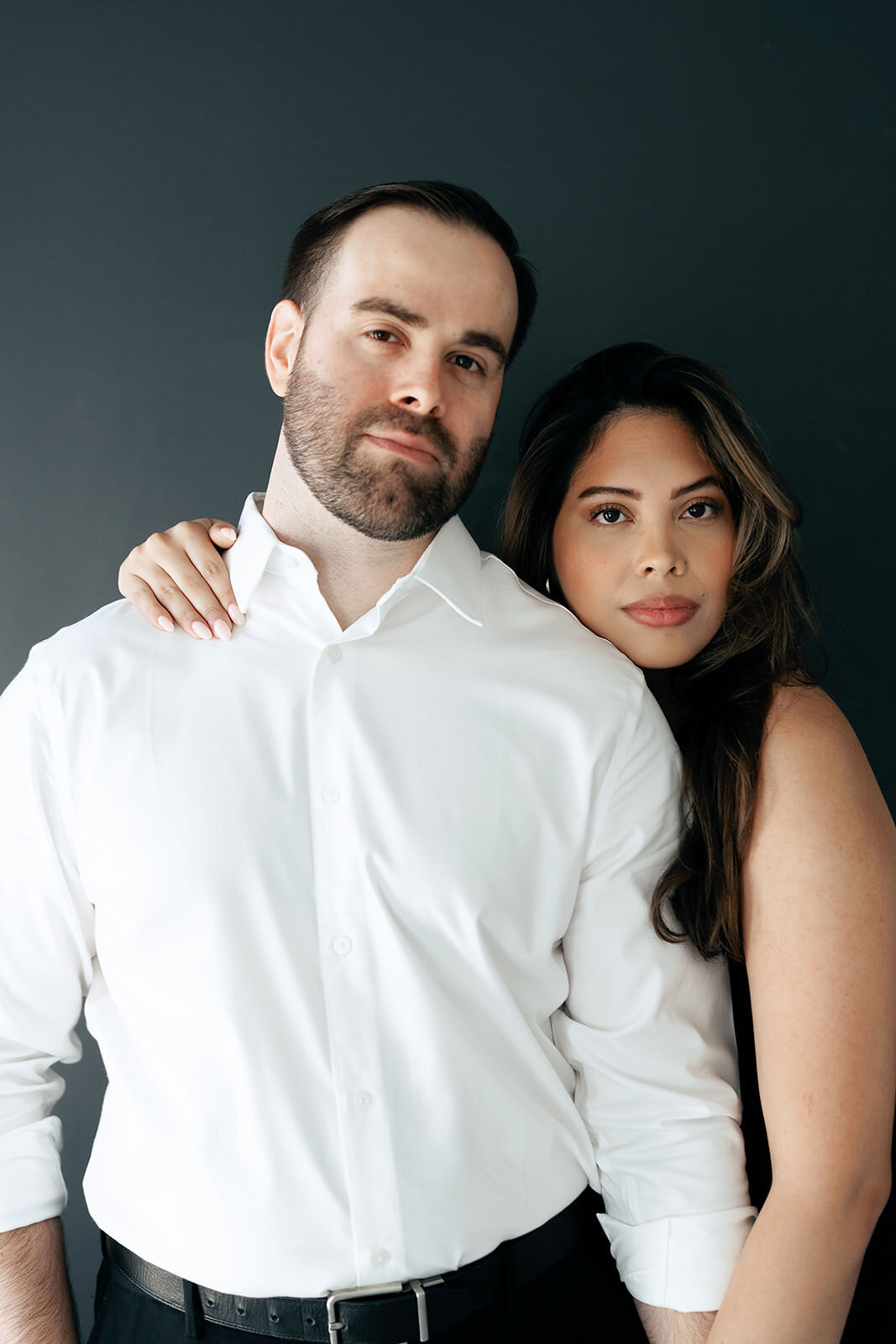 A couple pose together with serious faces against a black wall for their engagement photos at the Lumen room