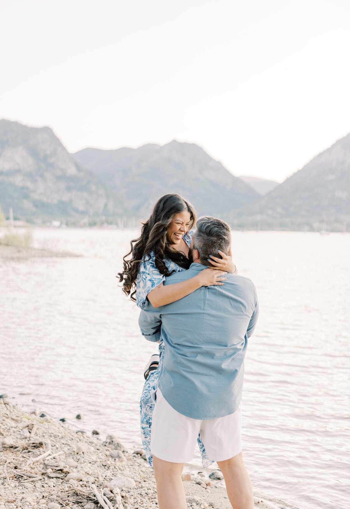 A man has his girlfriend wrapped in his arms. He has her lifted and is spinning her around while she smiles. They are on the shores of a lake. Taken by a local couples photographer.