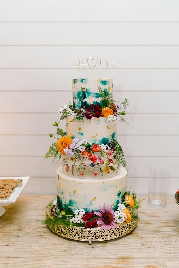 A four tier wedding cake sits on a gold cake stand. The cake is decorated with watercolor style teal accents, gold leaf, and an assortment of colorful fresh flowers around the bottom of each tier.