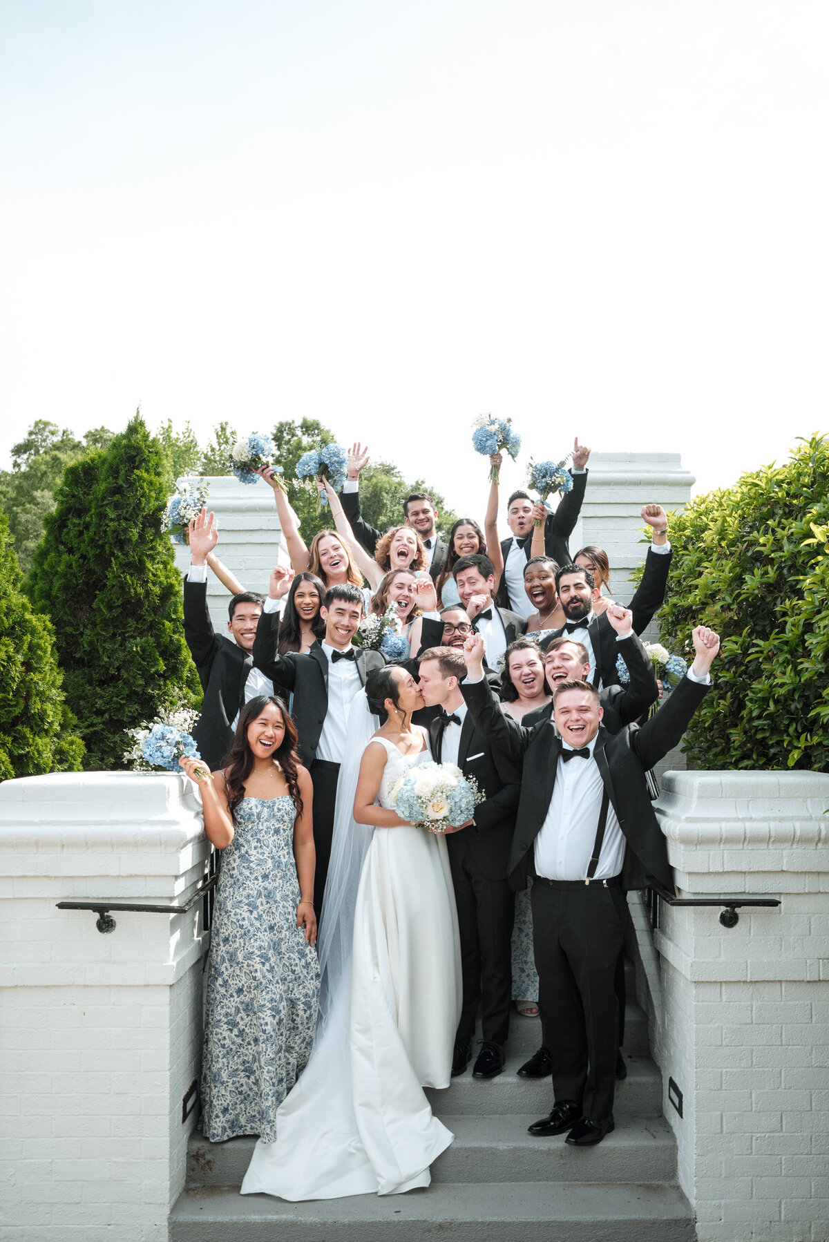 Wedding party celebration at Providence Country Club in Charlotte, NC. The bride and groom share a kiss while surrounded by cheering bridesmaids and groomsmen holding blue hydrangea bouquets. Captured by DeLong Photography, Charlotte wedding photographers specializing in luxury country club weddings and joyful candid moments.