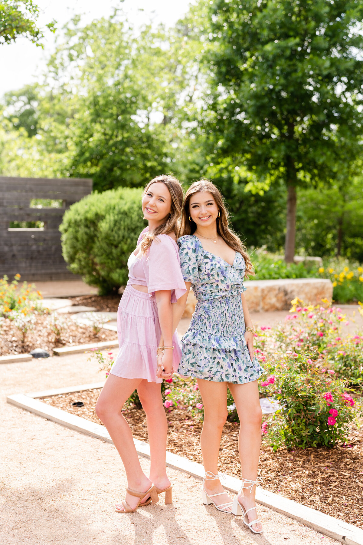 Texas A&M senior girls holding hands and back to back wearing pastel dresses in the middle of Leach Teaching Gardens