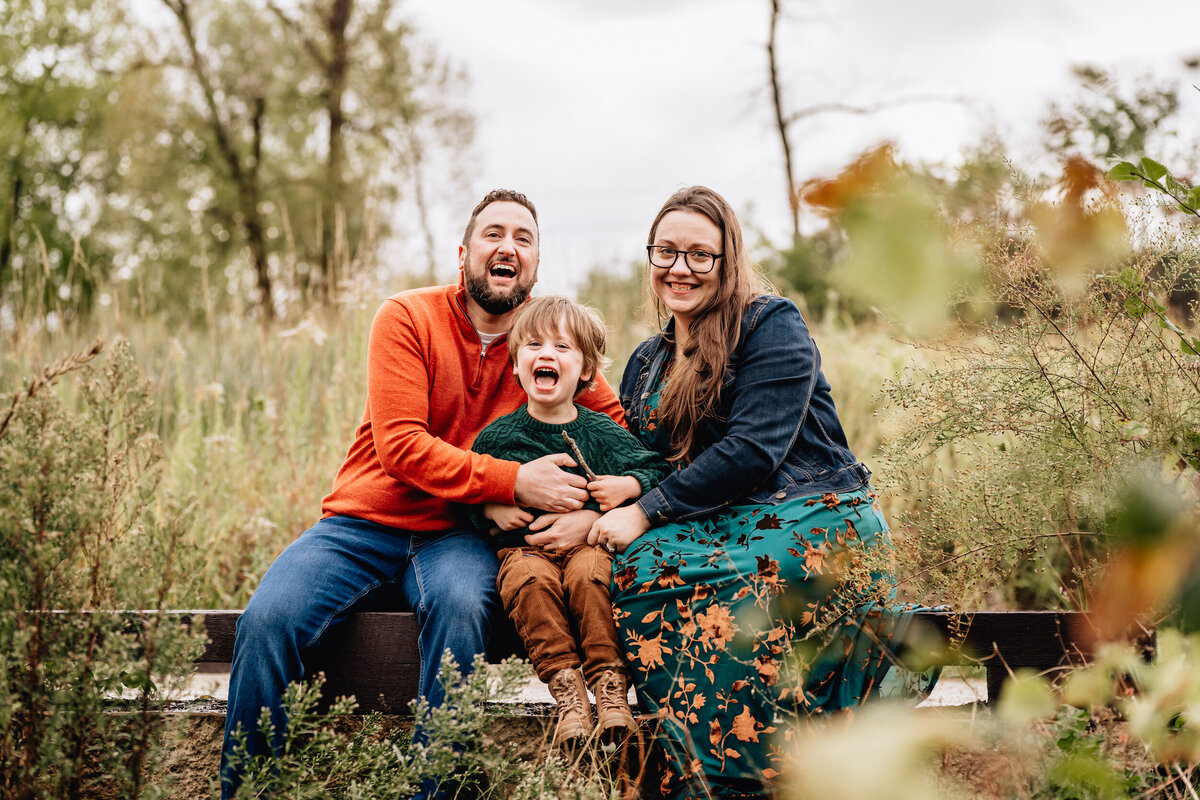 A family of three, with a young boy in the middle, sits on a wooden bench in a field. The boy is laughing joyfully while his parents, both smiling, sit on either side of him.