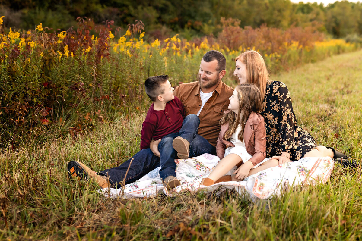 A family of four sits on a blanket in a grassy field, surrounded by wildflowers. Captured by a Bloomington family photographer, the parents look lovingly at their two children, a boy and a girl, who are smiling. The setting sun casts a warm glow over these incredible moments.