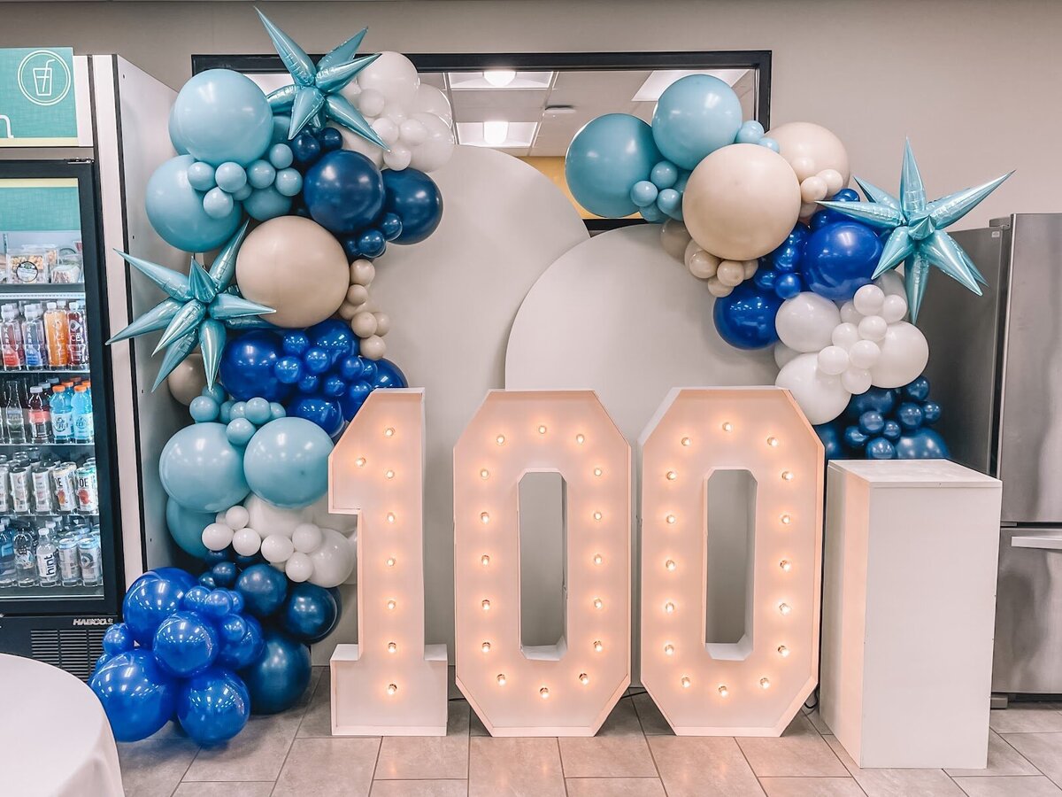 Blue and white balloon garlands attached to white wooden backdrops with giant marquee numbers for a 100th birthday party.