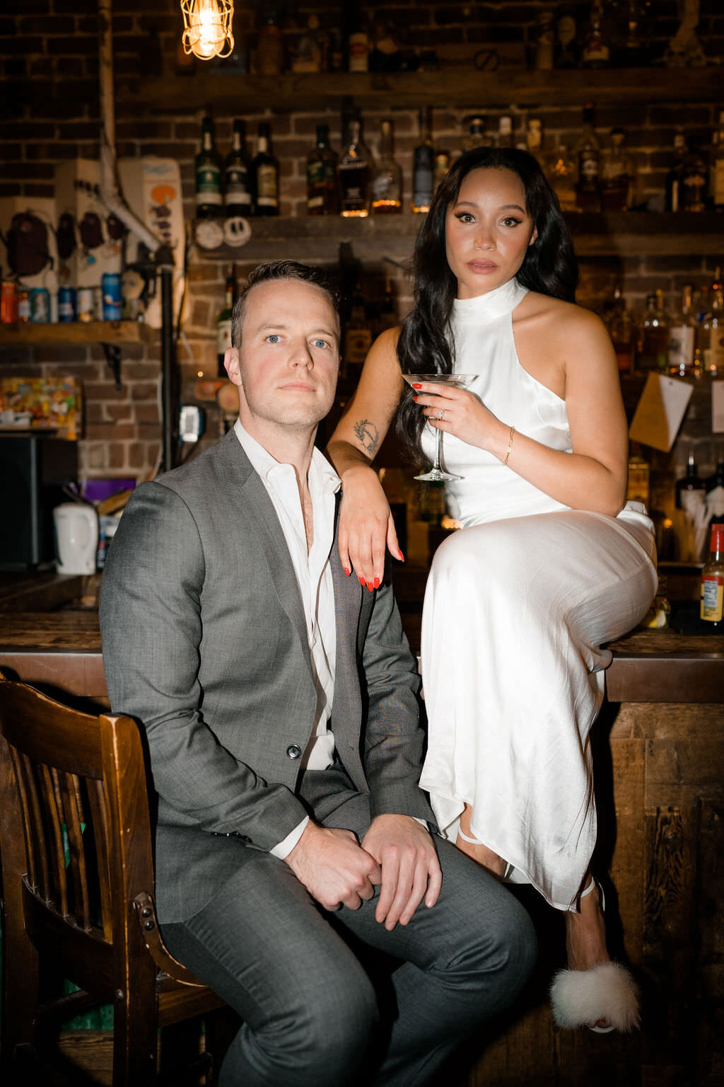 bride and groom posing at a bar while the bride sits on the bar holding a drink