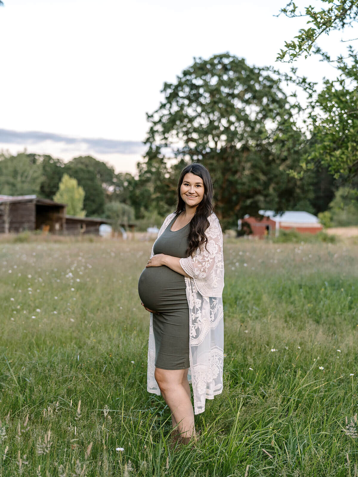 A woman stands outdoor with her hand on her pregnant belly and smiles at the camera.