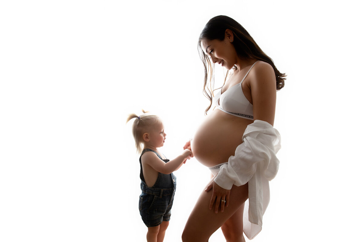 Pregnant mother wearing white underwear and a linen shirt fallen off her shoulders looking lovingly at her little girl who is touching her belly.
