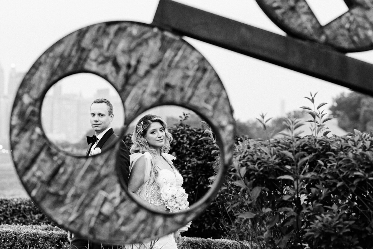 Bride and groom framed through a circular metal sculpture outdoors, looking toward the camera with the city skyline softly blurred behind them.