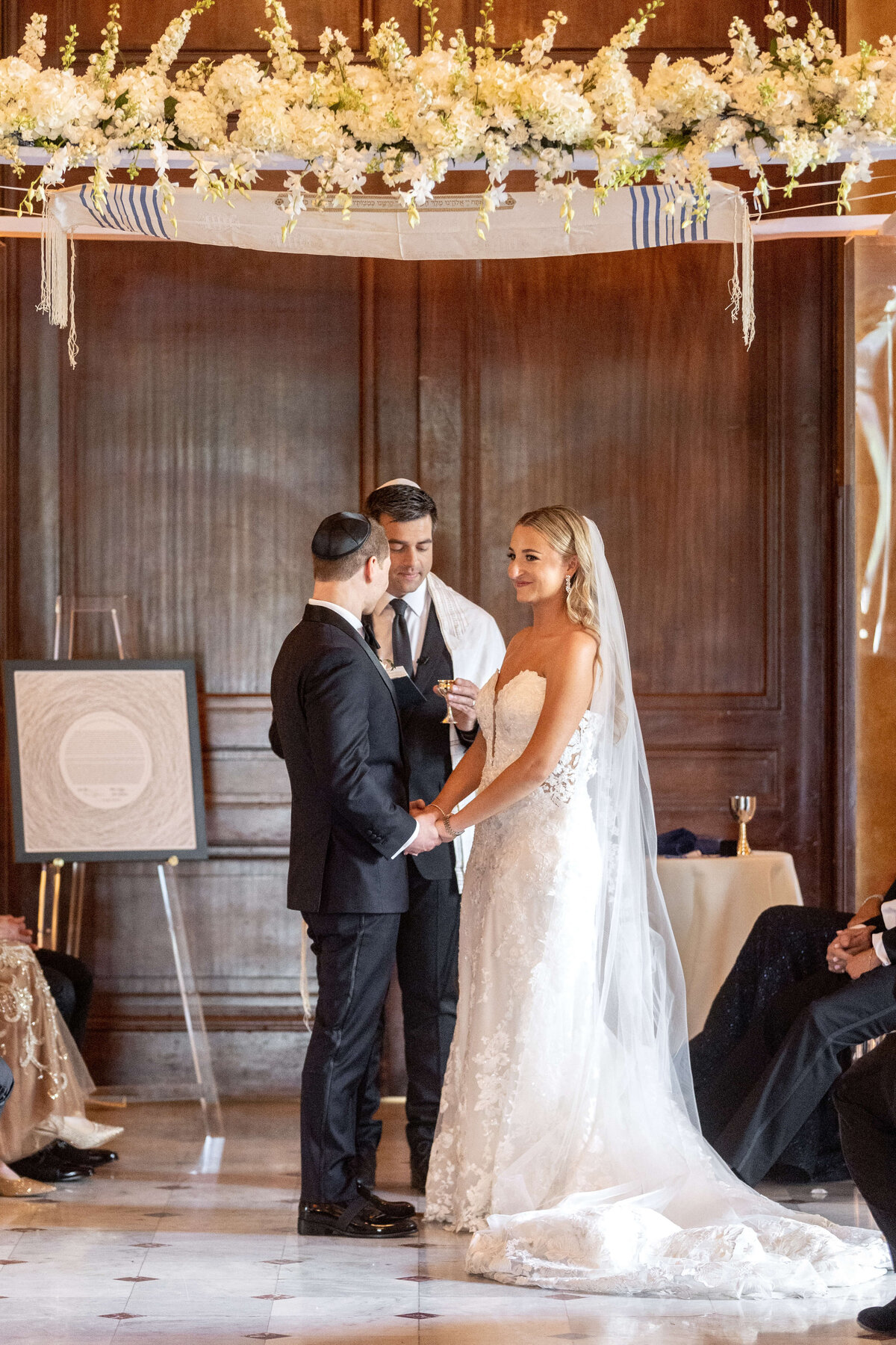 A bride and groom stand under a floral chuppah during their wedding ceremony, holding hands and exchanging vows. A man officiates the ceremony. The bride wears a white dress and veil, while the groom is in a suit with a kippah. Guests are seated on either side.