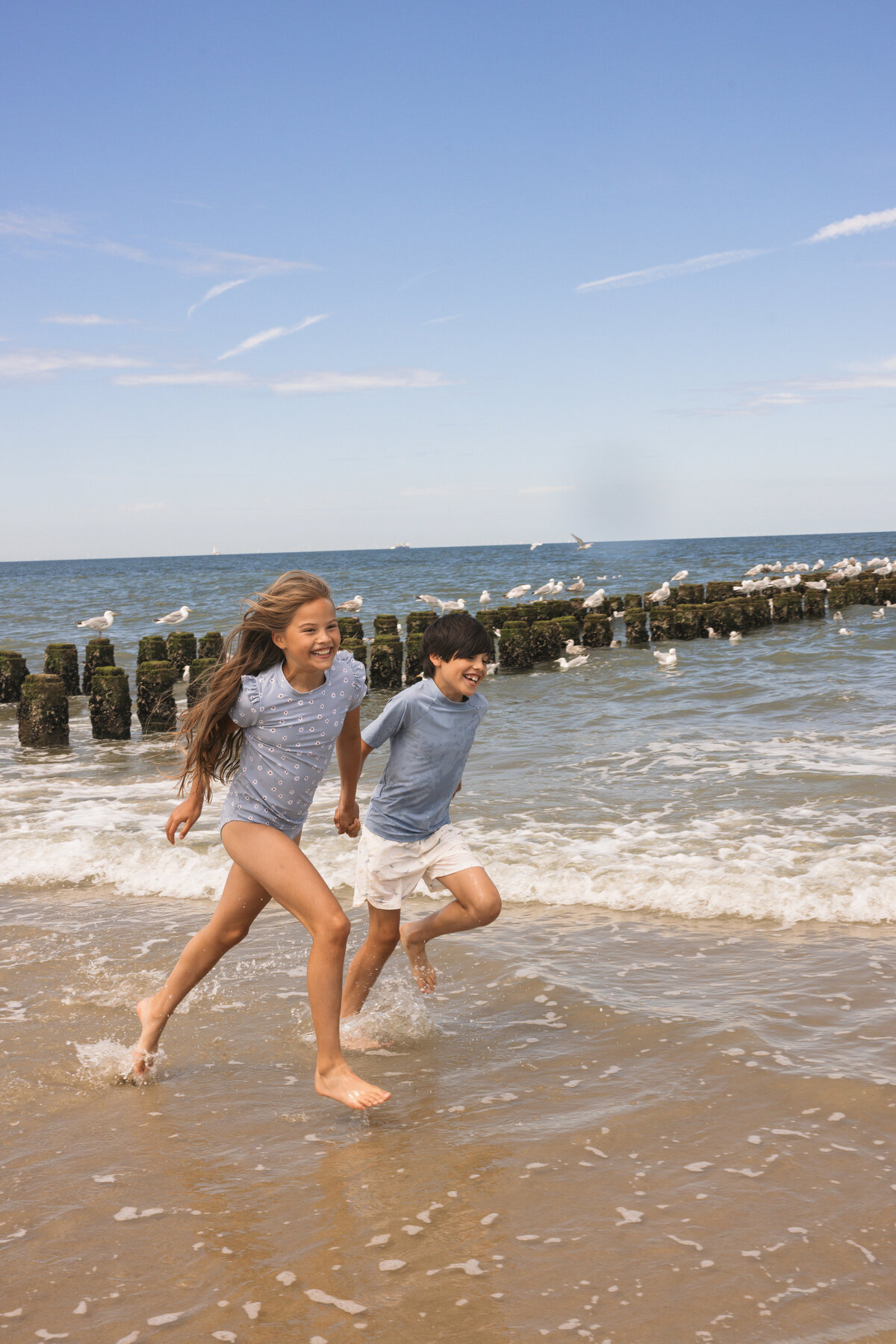 Two children laughing and running through the shallow waves, wearing Swim Essentials swimwear, with seagulls and breakwater posts in the background.