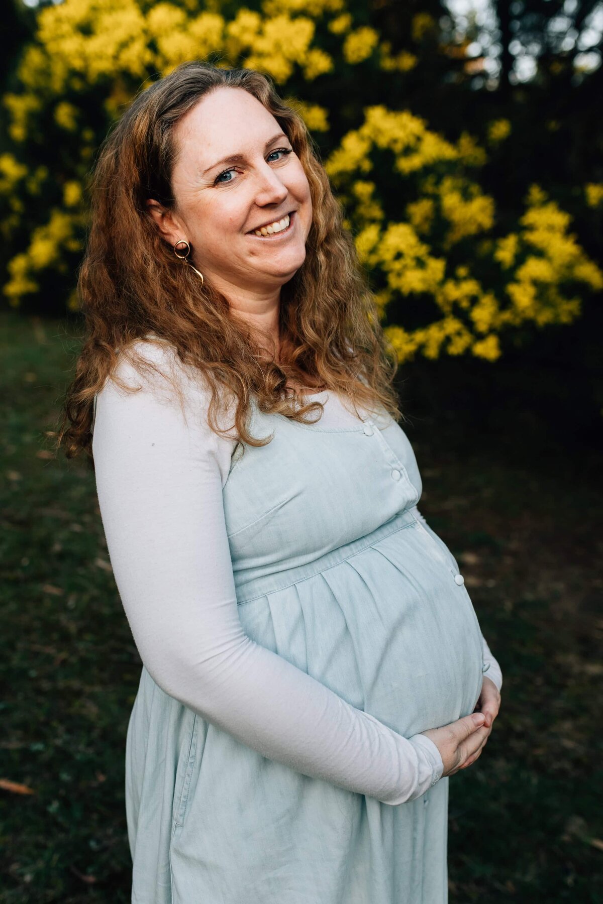 Pregnant woman in blue dress standing in front of wattle bush, Melbourne maternity photography.