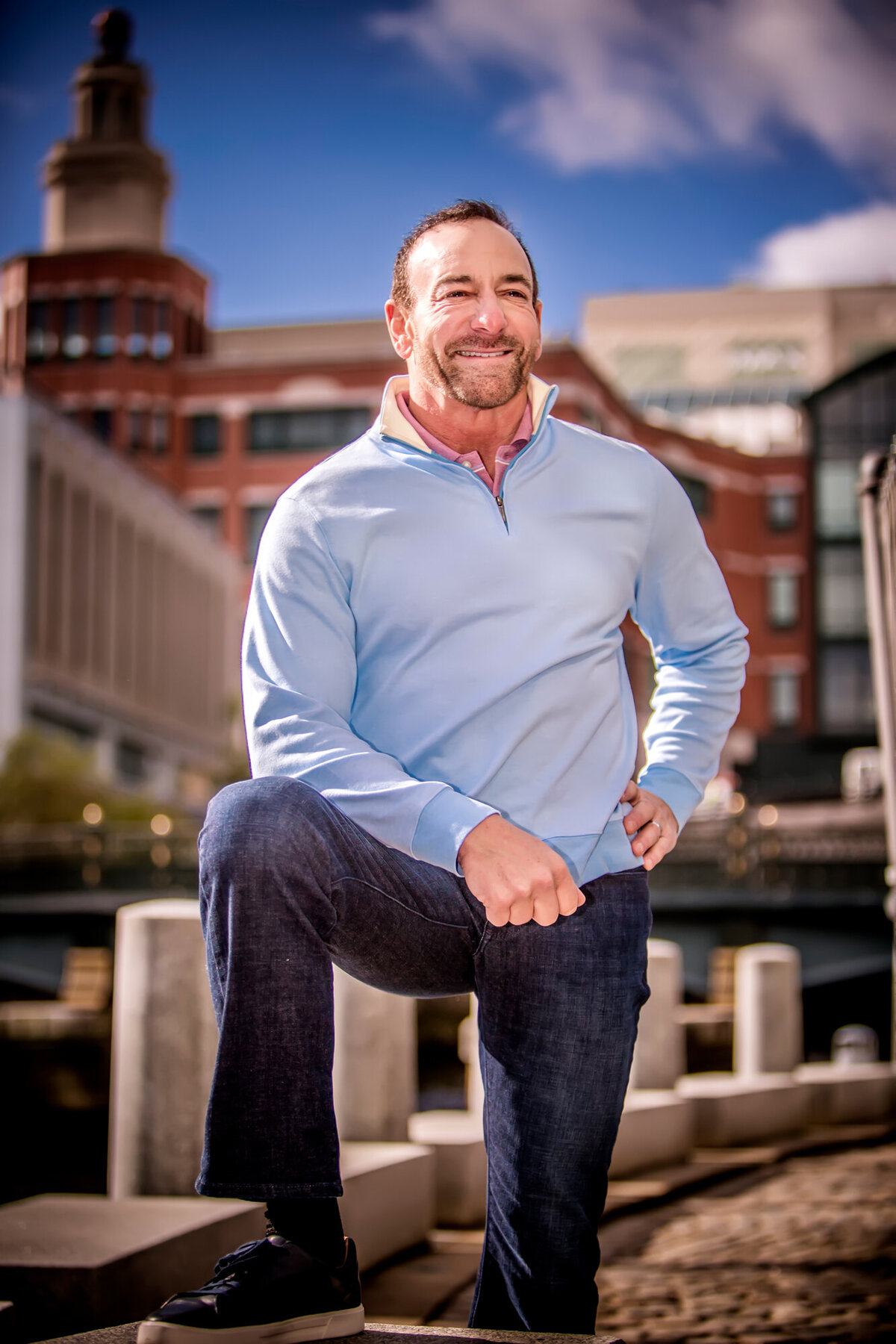 A male Providence doctor wearing a light blue sweater and jeans, standing outside with the city skyline behind him, while posing for a South Shore MA branding photographer