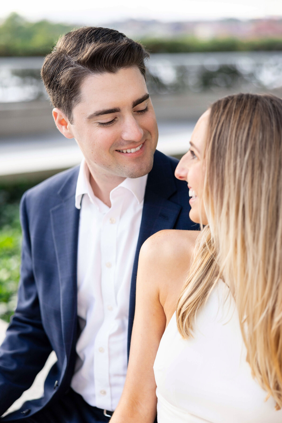 A couple sits together outdoors. The man, in a blue suit, smiles at the woman, who has long blonde hair and wears a white top. They appear happy and relaxed, with greenery and a blurred background behind them.
