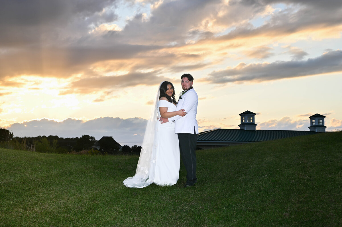 A beautiful wedding portrait of a couple embracing outdoors at sunset, with a scenic backdrop of a grassy hill, dramatic clouds, and soft golden light creating a romantic atmosphere.