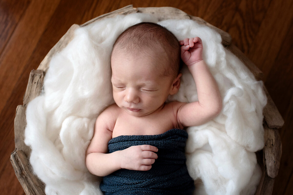 Newborn boy sleeping with one arm up and the other hand on his chest, laying in a round basket at his Allen, TX newborn photo shoot.