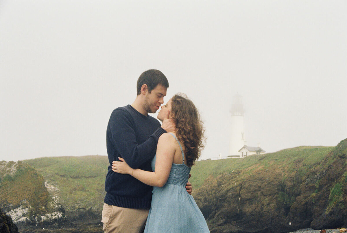 A couple embrace with a foggy lighthouse in the background, demonstrating engagement photos by Among the Oaks. 